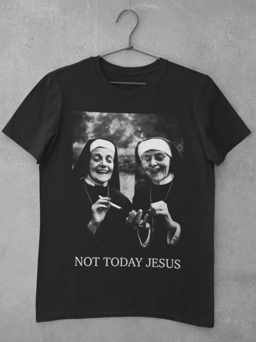Das Bild zeigt ein schwarzes T-Shirt mit zwei lachenden, rauchenden Nonnen als Motiv. Das T-Shirt ist auf einem Kleiderhaken aufgehängt. Es wird verwendet, um einen Eindruck zu vermitteln, wie das reale Produkt letztendlich aussehen wird.