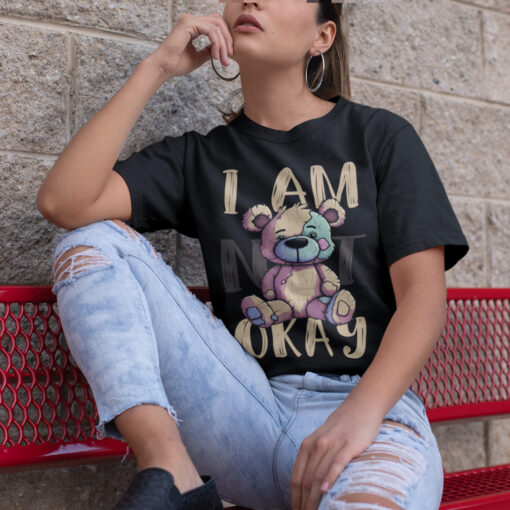 Das Bild zeigt eine Nahaufnahme einer Frau die ein schwarzes T-Shirt zum Thema psychische Gesundheit mit dem I Am (Not) Okay Design trägt. Es wird verwendet, um einen Eindruck zu vermitteln, wie das Produkt an einem Menschen aussieht.